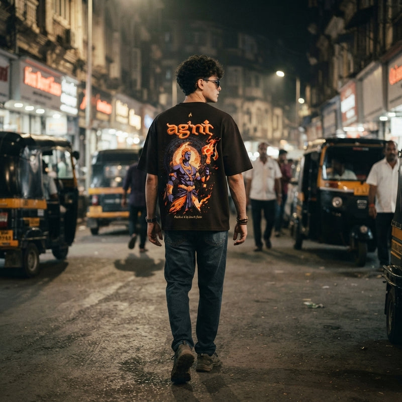 A young Indian man wearing a premium oversized puff-print T-shirt while walking down a bustling, dimly lit Mumbai street at n