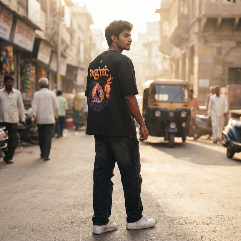 A young South Asian man wearing an Agni oversized puff print T-shirt walking down a sunlit city street.